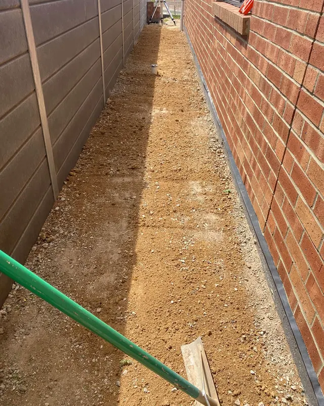 Dirt spread over crushed rock along a red brick wall, a rake handle at the left